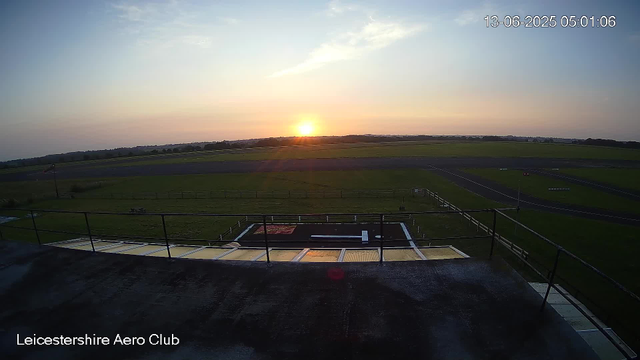 A sunrise over a grassy airfield with runways visible in the distance. The sky is partly cloudy, and the sun is near the horizon, casting a warm glow. There is a balcony or platform with a railing in the foreground, and the location is identified as Leicesterhire Aero Club. The timestamp shows 13-06-2025 05:01:06.