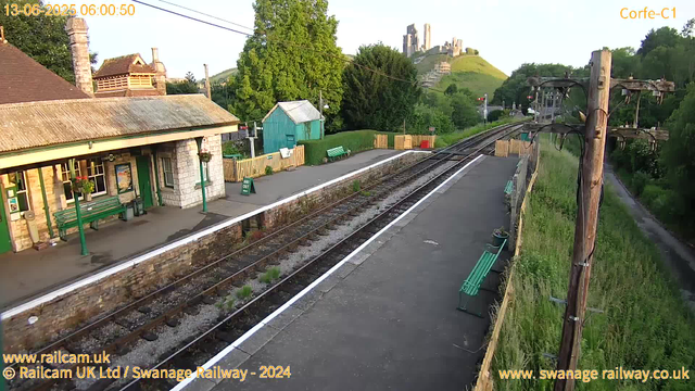 A small railway station platform is visible with a building on the left featuring green benches and flower baskets hanging from the roof. There are two signs on the platform, one indicating "Way Out." The railway tracks run through the center, with a wooden fence and green foliage along the right side, where a dirt path can be seen. A utility pole with wires is on the right side of the platform. In the background, a lush green hill with a castle or ruins sits atop, bathed in soft sunlight, indicating early morning. The timestamp in the upper left corner shows the date as June 13, 2025, at 6:00 AM.