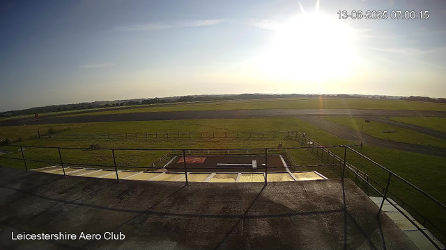 An outdoor view taken from a webcam at Leicestershire Aero Club shows a sunlit sky with the sun near the horizon on June 13, 2025, at 07:00:15. In the foreground, there is a fenced area with what appears to be a small aircraft or equipment on a platform. Beyond that, there are open grassy fields with some paved runways or taxiways, stretching into the distance under a clear sky. The overall scene suggests a quiet, early morning at an aviation club.
