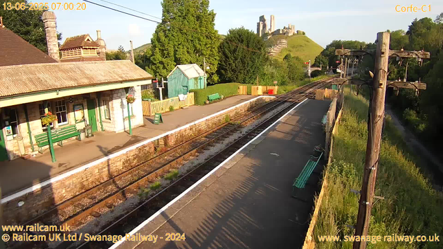 A small railway station platform with a stone building on the left, decorated with flower baskets and green benches. The platform is partly shaded by a utility pole with overhead electrical wires. In the background, there is a lush green hill with a castle ruin on top, and trees surrounding the area. The sky is clear, and the sunlight suggests early morning. The image also includes some wooden barriers near the edge of the platform.