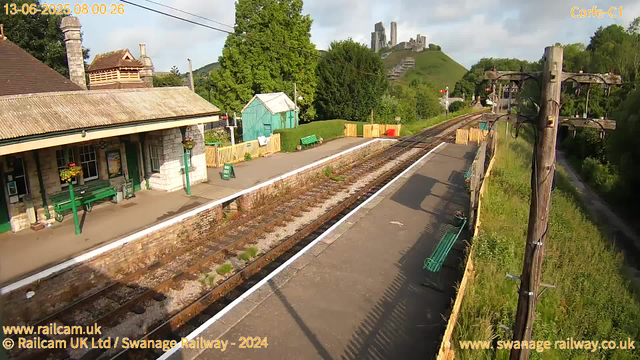 A small railway station platform with a stone building featuring a tiled roof and green benches. There are flower baskets hanging from the building. Green trees and a small turquoise shed are visible in the background. A railway track runs parallel to the platform, with a few plants growing between the sleepers. To the right, there are wooden utility poles with wires. In the distance, a hill with ruins or a castle on top is partially covered by trees. The sky is partly cloudy, and the overall scene is bathed in sunlight.
