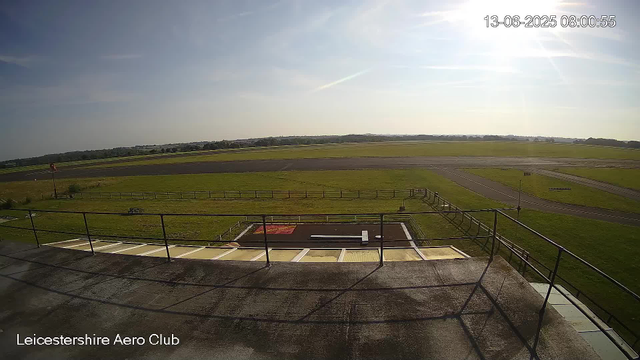 An outdoor view from a webcam at Leicestershire Aero Club showing a bright, sunny day with clear skies. In the foreground, there is a balcony with a metal railing and some parked vehicles. Beyond the balcony, there is a fenced grassy area, a small tarmac platform, and a runway extending into the distance. The landscape is flat, with open fields and a treeline on the horizon. The timestamp on the top right indicates the date and time as June 13, 2025, at 8:00 AM.