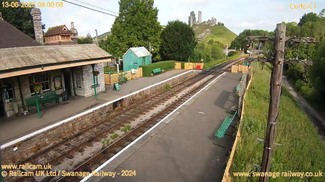 A small railway station platform with a stone building on the left, featuring a tiled roof, a chimney, and hanging flower baskets. There are green benches and a green shed further along the platform. The railway tracks run through the center, with some grass and weeds growing between the rails. To the right of the tracks, a wooden fence with yellow and natural-colored sections separates the platform from a grassy area. In the background, there are trees, a hill or small mountain with a medieval castle ruin on top, and distant green hills. An overhead electrical line pole with wires extends along the right side of the image. The top left corner of the image shows the date and time as June 13, 2025, 09:00:08.
