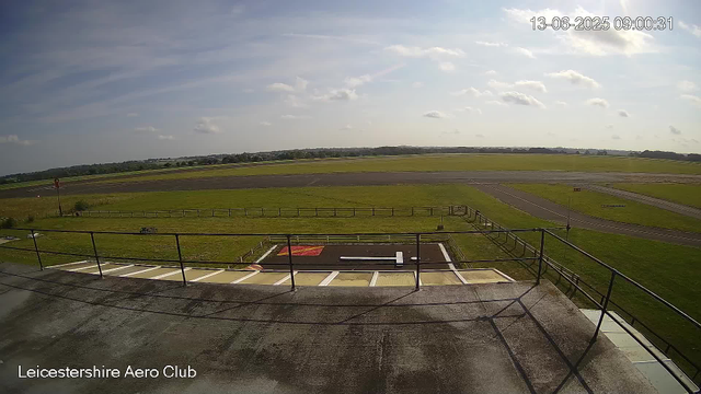 An outdoor scene at Leicesterhire Aero Club showing a runway and grassy fields under a partly cloudy sky. The view is from a balcony or observation deck with a railing, and there are painted lines and parking spaces on the concrete surface in the foreground. The landscape extends to the horizon with distant trees and structures visible. The time and date are displayed in the top right corner: 13-06-2025 09:00:31.