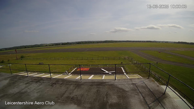 A view from a webcam at LeicesterShire Aero Club showing a grassy airfield with taxiways and open sky. The date and time displayed in the top right corner is 13-06-2025 10:00:03. The foreground includes a section of the tarmac with painted markings and a railing, while the background consists of flat grass fields extending to the horizon under a partly cloudy sky.