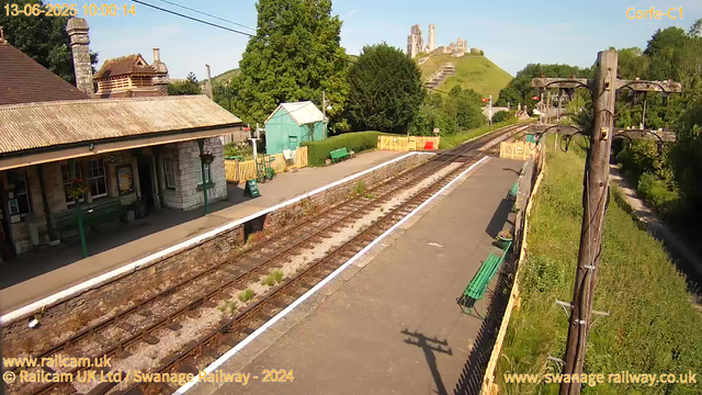 A small rural train station with a single platform, railway tracks running along the center, and a station building on the left side. There are green benches on the platform, and a pathway on the right side lined with a wooden fence and greenery. A telephone pole with electric wires stands near the platform. In the background, there is a lush green hill with the ruins of an ancient castle or fortress atop it. The sky is clear and bright, indicating a sunny day.