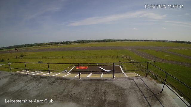 A wide-angle view of a small aircraft parking area at Leicestershire Aero Club, with a concrete platform and metal railing in the foreground. A marked helicopter landing pad with a white 'H' and red markings is visible. In the background, an open grassy airfield stretches out under a partly cloudy sky, with distant trees on the horizon. The timestamp shows the date as June 13, 2025, at 11:00 am.