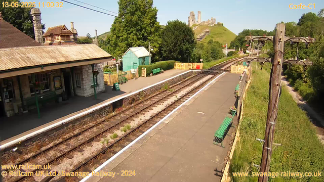 A small railway station platform with a stone building on the left side, featuring a chimney and a metal roof. There are green benches and potted plants along the platform. Railway tracks run parallel to the platform, with a lush green landscape and trees surrounding the area. In the distance, a hill with the ruins of a castle or old fortification is visible under a clear blue sky. On the right, there are electric poles and a narrow grassy strip. The timestamp shows the date as June 13, 2025, and the time as 11:00:32.