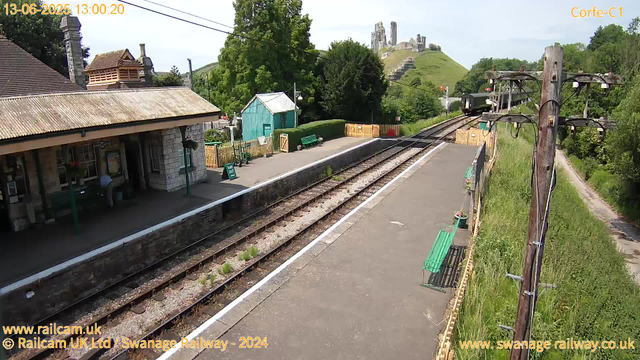 A small train station platform with a building on the left, benches, and a sign that says "Way Out." A train is approaching in the distance on the tracks, which are bordered by a yellow fence and surrounded by greenery. There are trees, a hill with ruins or an old structure in the background, wooden utility poles with wires, and a dirt path running alongside the station. The sky is mostly clear with some light clouds. The timestamp on the image shows "13-06-2025 13:00:20."