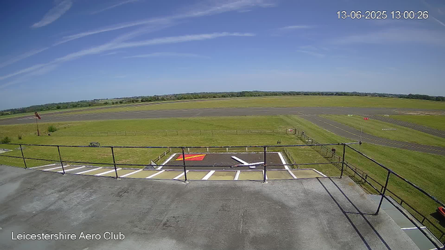 An outdoor view from a webcam at Leicestershire Aero Club shows a clear sky with a few wispy clouds and a vast open grassy area. The foreground includes a concrete platform with a railing, some painted markings, and a small red painted area. There is a windsock leaning slightly, and a single person is visible walking along a pathway. In the background, there are sections of a runway or taxiway with painted markings, surrounded by green fields and distant trees under bright daylight. The top right corner displays a timestamp: 13-06-2025 13:00:26.