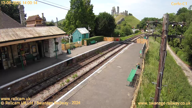 A small train station platform with a building on the left, featuring a person standing near the entrance. There are empty green benches along the platform, and a small turquoise shed in the background. Railroad tracks run through the middle of the scene, with a grassy area and trees on the right side. In the distance, a hill with the ruins of an ancient castle or fortress is visible atop it. The sky is mostly clear with some light clouds. The timestamp indicates the photo was taken on June 13, 2025, at 14:00.