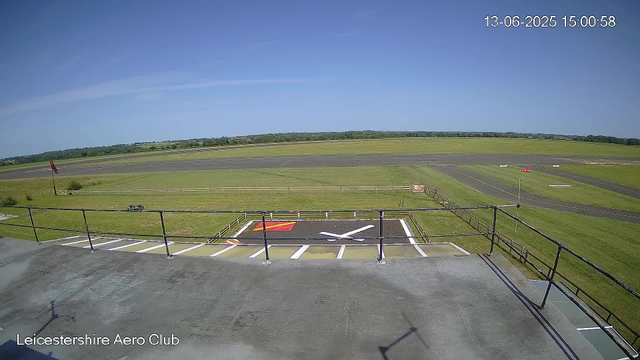 An empty aircraft parking area at Leicestershire Aero Club, with a helipad in the foreground marked with a large white "H" and red and yellow painted areas. The scene is open with a grassy airfield extending into the distance under a clear blue sky. The timestamp in the upper right corner shows "13-06-2025 15:00:58".