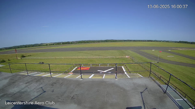 An empty tarmac area with a grassy perimeter, a few painted markings, and a small fenced section with a red and white arrow. The background features an open field under a clear blue sky, with a distant tree line. The image is marked "Leicestershire Aero Club" at the bottom left corner, and the top right corner shows a timestamp: 13-06-2025 16:00:37.