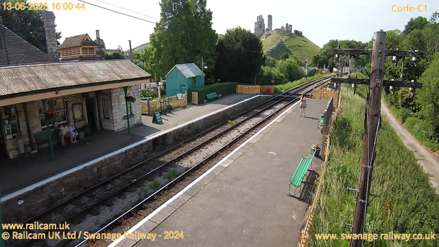 A small train station platform with a building on the left side, two people sitting on benches under a covered area, and one person standing further down the platform. There are railway tracks running parallel to the platform, with wooden fencing and green benches along the side. In the background, there are lush green trees, a small blue wooden shed, and a hill with ruins of an ancient castle or fort at the top. There is also a utility pole with wires running across the scene, and potted flowers on the benches. The sky is clear and sunny. The timestamp shows the date as June 13, 2025, at 4:00 PM.