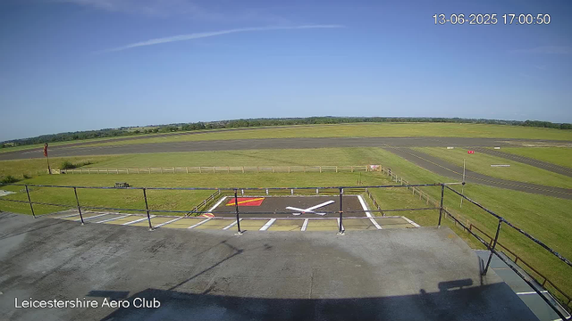 A clear day at Leicesterhire Aero Club, with a view of the runway and surrounding fields. The sky is bright blue with a few wispy clouds, and there are no aircraft visible on the runway or in the sky. The image is timestamped June 13, 2025, at 5:00 PM.