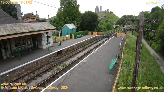 A small rural train station platform with a stone building on the left, decorated with hanging flower baskets and a bench. The platform is bordered by a white line, with a "Way Out" sign near the edge. There are two green benches, one on the platform and one further back, and some potted plants. On the right side, there is a wooden utility pole with electrical wires. In the background, green hills and trees are visible, with an old stone castle ruin perched atop the hill. The scene appears to be during late afternoon or early evening, as indicated by the timestamp.