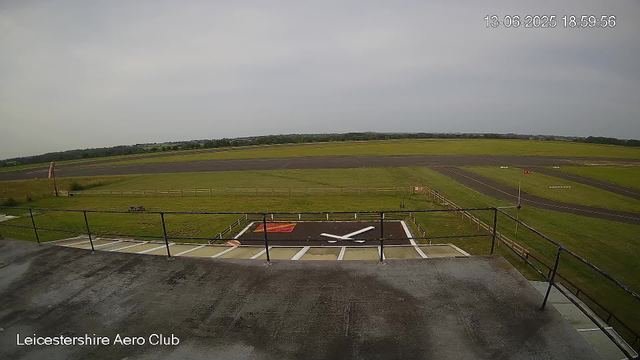 An empty airplane parking area with a helicopter landing pad in the foreground. The landing pad has a large white "H" and a red square. There is a black railing around the edge of the top terrace from which the photo is taken. Beyond the landing pad, there is a grassy area with a flagpole on the left side. In the distance, there are taxiways and runways, and an open field stretching to the horizon under a partly cloudy sky. The timestamp on the top right indicates the date and time as 13-06-2025, 18:59:56. The bottom left corner shows the location as Leicestershire Aero Club.
