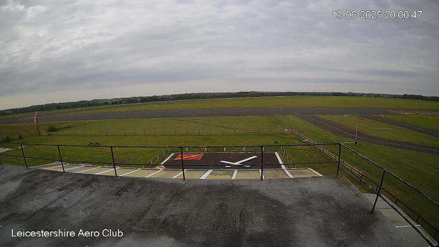 A wide view of the Leicestershire Aero Club's airfield shows an overcast sky with gray clouds. In the foreground, there's a rooftop balcony with a black railing, and a large X-shaped marking is painted on the flat surface below. The airfield features an open grassy area with a small, fenced-in section and a paved runway extending into the distance. In the background, the landscape is flat with some trees and distant buildings. The timestamp on the top right indicates it was taken on June 13, 2025, at 8:00 PM.