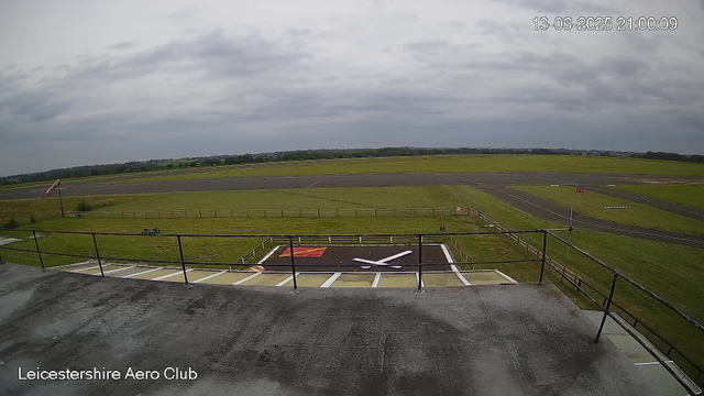 An empty helicopter landing pad with a red and white cross symbol, surrounded by safety railings, at Leicestershire Aero Club. Beyond the pad, there is a grassy field and an airstrip extending into the distance, under a cloudy sky. The timestamp in the top right corner indicates the date and time as 13-06-2025 at 21:00:09.