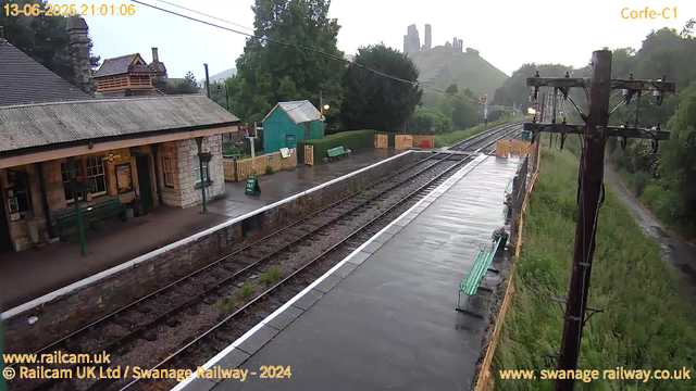 A wet railway station platform in a rural area during early evening, with a small stone station building on the left, green benches, a wooden fence, and a green shed in the background. Overhead electric wires stretch across the platform, and a utility pole stands on the right. The sky appears overcast, and distant hills with ruins on top are visible in the background. The scene looks peaceful and quiet.
