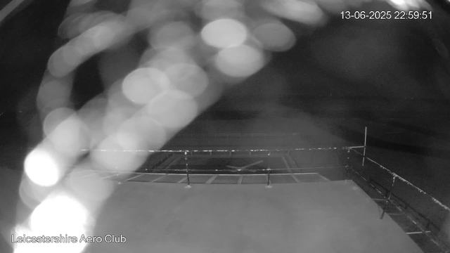 A black-and-white image of an empty tennis court at night, with a fence surrounding it. The court appears to be wet or reflective, and there are some bright bokeh light reflections in the upper left part of the image. The timestamp in the top right corner shows the date as 13-06-2025 and the time as 22:59:51. The bottom left corner has the text "Leicestershire Aero Club."