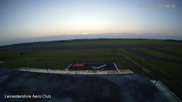 An early morning view at Leicestershire Aero Club showing a runway under a clear sky with the sun just beginning to rise on the horizon. The image includes a helipad in the foreground, with a marking of a white helicopter silhouette in a black box. The timestamp indicates the photo was taken on June 14, 2025, at 5:00 AM.