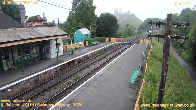 A small railway station platform with a stone building on the left, green benches, and a green fence. There are two train tracks running through the middle, with some grass and weeds growing between the sleepers. On the right side, there's a tall wooden utility pole with wires, a narrow gravel path alongside the tracks, and an area of lush greenery and trees. In the background, a hill with a cityscape or castle ruins is visible, shrouded in light mist. The image has a timestamp of 14-06-2025 05:00:42.