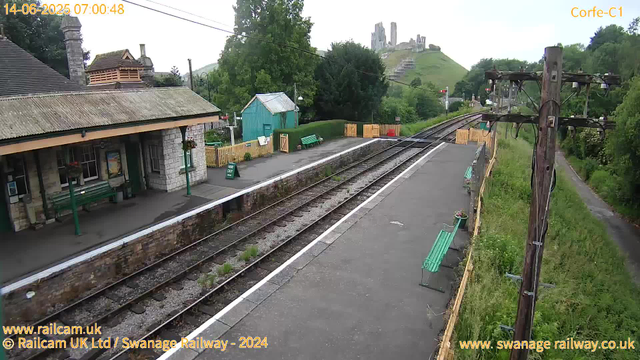 A small, rural train station platform with a stone building on the left, featuring a chimney and a small overhang. There are green benches, a green "Way Out" sign, and some flower baskets hanging from the building. A blue shed is behind the benches. The railway tracks run through the center, with some greenery growing between the sleepers. To the right, there is a wooden utility pole with electrical wires. In the background, a green hillside with a castle ruin on top is visible, along with trees and a pathway on the right side of the image. The timestamp indicates early morning on June 14, 2025.