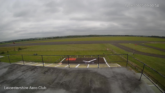 The image shows a view from a webcam at Leicesterhire Aero Club, capturing a cloudy sky over an airfield. In the foreground, there is a helicopter landing pad with a red and yellow marking and a white rotor blade symbol. A small grassy area with a bench and a windsock on a tall pole are visible to the left. The runway stretches into the distance, with a few markings and signs visible along its length. The landscape beyond the airfield is flat with distant trees and fields under a predominantly overcast sky. The timestamp on the top right reads 14-06-2025 09:00:54.