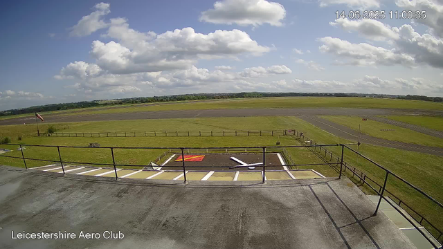 A wide view of an airport runway and surrounding grassy fields under a partly cloudy sky. The foreground shows a parking area with marked parking spots, a railing, and a large white arrow painted on the ground pointing to the right. There is also a red square with a yellow border and a yellow arrow next to it. The timestamp in the upper right corner reads "14-06-2025 11:00:35," and the bottom-left corner credits "Leicestershire Aero Club."