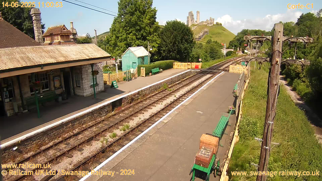 A small train station platform with green benches, a small green shed, and a stone building with a tiled roof. Railroad tracks run through the scene, and there are lush trees and a grassy hill with a castle ruin on top in the background. A wooden utility pole with wires is on the right side of the image. The weather is sunny with a clear sky, and the date and time are displayed at the top left corner.