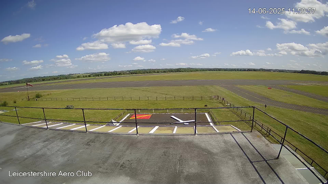 A view from a webcam at Leicestershire Aero Club shows a clear day with a bright blue sky scattered with fluffy white clouds. In the foreground, there is a parking area with white painted lines, and one marked with a red "H" for helicopter landing. A black railing runs along the edge of the parking area. Beyond that, there is a grassy field leading to a paved taxiway and runway, with a windsock on the left side. The background features an open landscape with trees and fields. The top right corner displays the date and time: 14-06-2025 11:59:57.
