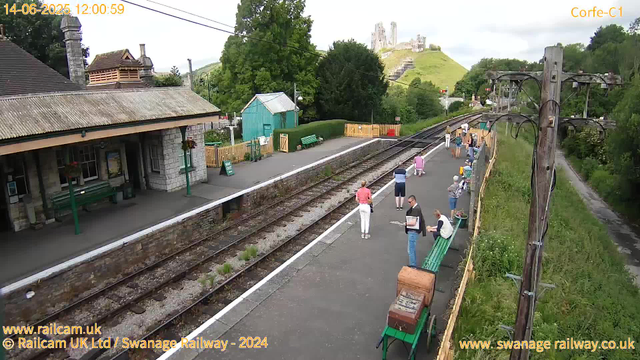 A small train station platform with several people waiting. There are benches, some people sitting and others standing, some reading, and some walking. The platform is bordered by a yellow fence on one side, with greenery and a path beyond. On the other side, there is a station building with stone walls and a corrugated roof, and a small green shed nearby. In the background, a hill with a historic castle ruins is visible atop, surrounded by trees. Utility poles with electrical wires run along the right side of the image. The timestamp shows it is around noon on June 14, 2025.