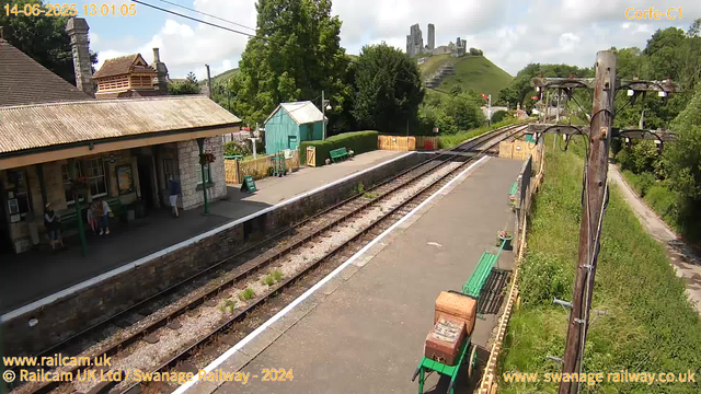 A small railway station platform with a few people waiting, surrounded by greenery and trees. There are two green benches on the platform, with one person sitting and a small child standing next to them. An old wooden utility pole with wires extends along the right side of the platform. Behind the station, a turquoise shed and a wooden fence are visible. In the background, on a hill, there are ruins of an ancient castle or fortress overlooking the landscape. The sky is partly cloudy, with patches of blue visible. The date and time in the corner indicate June 14, 2025, at 1:01 PM.