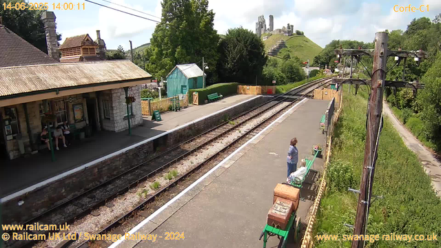 A small train station platform with two women sitting on a bench and one woman standing nearby. The station building is on the left, with a few people sitting under its shelter. The platform is bordered by railway tracks, and there are some green benches and flower pots along its edge. In the background, there is a hill with ruins of an old castle on top, and lush green trees surround the area. Electricity poles run along the side of the tracks, and the scene appears to be bright and sunny. The timestamp indicates the photo was taken on June 14, 2025, at 2:00 pm.