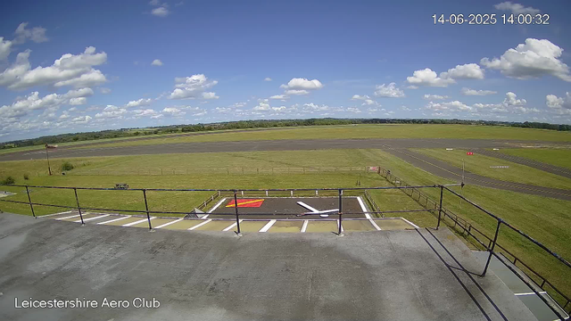 A clear sky with scattered white clouds over an empty airfield runways and green fields in the distance. The photo appears to be taken from a webcam at Leicesterhire Aero Club, as indicated by text at the bottom left corner. The date and time displayed are 14th June 2025 at 14:00:32. In the foreground, there is a parking area with some markings and a small fence.