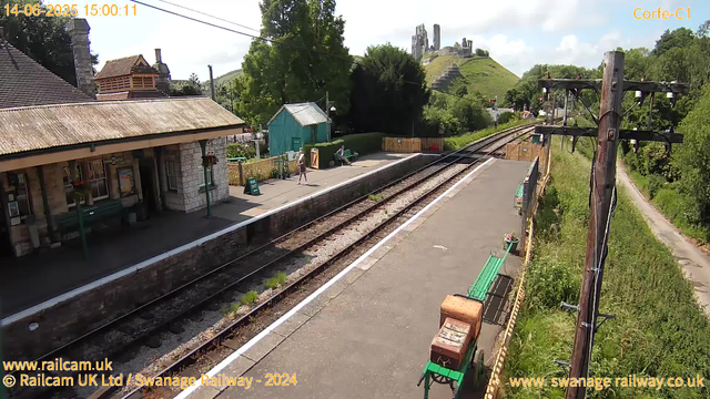 A small train station platform with a single passenger walking along the edge. There are green benches, one with a person sitting, and a small teal-colored building behind a wooden fence. Railway tracks run parallel to the platform, with a power pole on the right side. In the background, there is lush greenery and a hill topped with castle ruins. The sky is partly cloudy and bright.