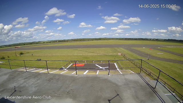 A view of an airfield under a partly cloudy sky with white fluffy clouds. In the foreground, there is a balcony with a railing, and a designated parking area with white lines and a marked helipad. The runway stretches across the middle distance, with green grass on either side. The landscape is flat with some distant trees on the horizon. The image includes a timestamp in the upper right corner showing "14-06-2025 15:00:06" and the text "Leicestershire Aero Club" at the bottom left corner.