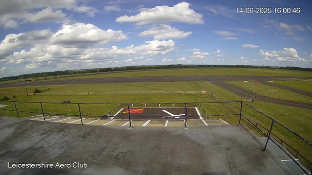 A view from a webcam at Leicesterhire Aero Club showing a sunny sky with scattered clouds over an airfield. In the foreground, there is a small helipad with a white helicopter symbol, colored red and yellow markings, and a parking area with painted lines. Fencing surrounds the helipad, and an open grassy area extends across the middle of the scene. In the distance, there are runways, grassy fields, and a landscape with trees and low hills under a bright sky with fluffy clouds. The timestamp in the upper right corner reads "14-06-2025 16:00:46."
