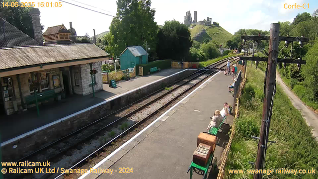 A small train station platform with a few people waiting, some sitting on benches and others standing. There are a couple of utility poles with wires overhead, lush greenery surrounding the platform, and a hill with ruins or an old structure visible in the background. The weather appears sunny with clear skies, and the scene captures a peaceful rural setting.