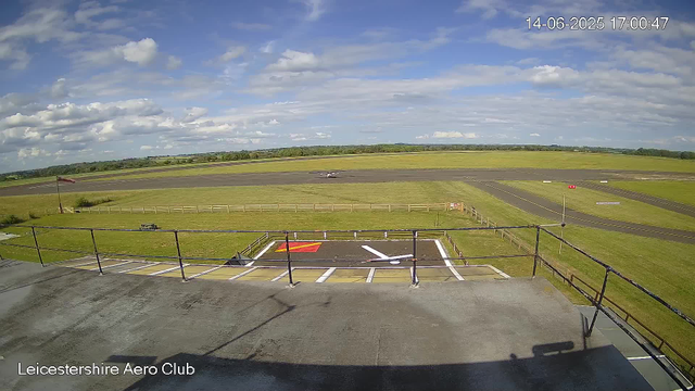 A view from a webcam at Leicestershire Aero Club shows a runway under a partly cloudy sky with patches of blue. In the foreground, there is a small parking area with space marked for vehicles, and on the left, a flagpole with a windsock. The grassy area surrounds the runway with some signs and markers. The horizon line reveals a gently rolling landscape with trees and fields in the distance. The date and time stamp on the top right corner reads 14-06-2025 17:00:47.