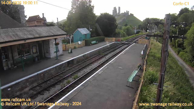 A small railway station platform with a stone building on the left, several green benches, and a small blue shed in the background. The train tracks run centrally, curving slightly to the right, with some overgrown grass between the rails. To the right, there are wooden utility poles with wires hanging between them. In the distance, a hill with ruins or an old castle overlooks the scene. The time and date are displayed at the top left corner: June 14, 2025, 19:00:11.