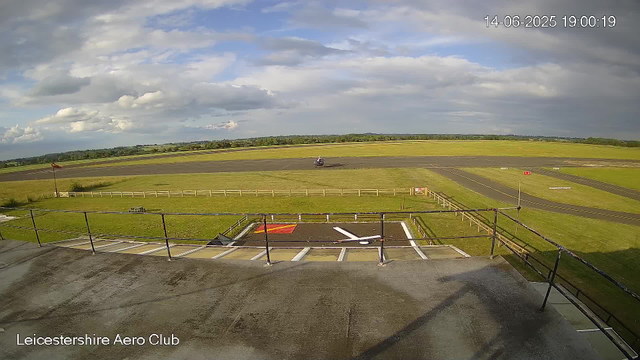 A view from a webcam at Leicestershire Aero Club shows an open grassy area with a runway and a helicopter taxiing on the ground. In the foreground, there is a raised platform with a black railing, part of which has parking marks and a red and white symbol. The sky is partly cloudy with patches of blue, and the timestamp indicates the date as June 14, 2025, at 7:00 PM.