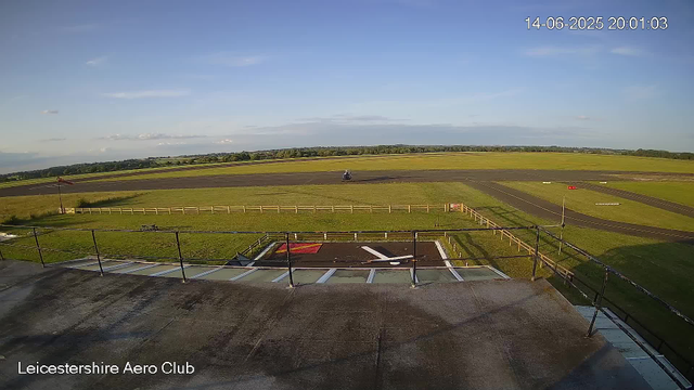 A view from a webcam at Leicestershire Aero Club showing the runway and tarmac area in bright daylight. In the distance, there is a helicopter on the grass. The sky is mostly clear with a few clouds, and the timestamp indicates it is June 14, 2025, at 8:01 PM.