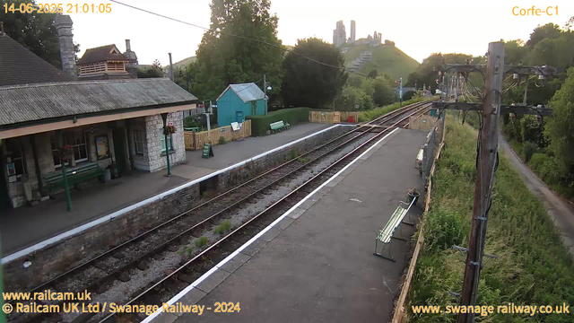 This image shows a peaceful train station platform at dusk with a small stone building on the left, topped with a tiled roof. The platform has green benches and some flower pots hanging from lamp posts. There are three railway tracks running parallel, with some weeds growing between the rails. On the right, there is a line of electrical poles with wires overhead and a narrow dirt path running alongside the plants and foliage. In the background, there are green hills with trees and a hilltop castle or ruins. The sky is fading into evening light. The image is marked with a timestamp in the top left, and URLs and credits are displayed at the bottom.