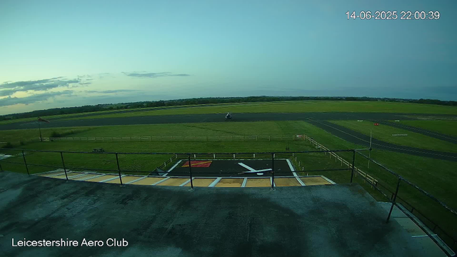 An aerial view from a webcam at Leicesterhire Aero Club shows a small aircraft on a grassy field adjacent to a runway. The sky is partly cloudy with a few clouds near the horizon, and the time stamp indicates it is 10:00 PM on June 14, 2025. The scene is calm with no signs of motion or activity.