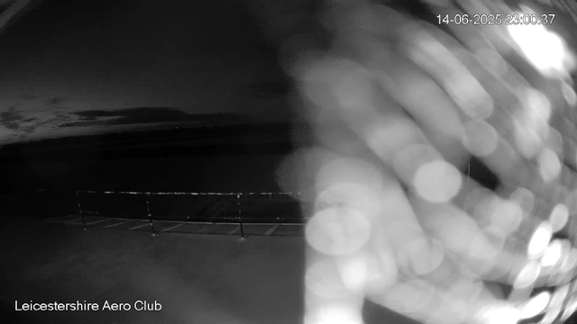 A black-and-white webcam image shows an empty outdoor area at night with a sports fence or goalpost and open landscape in the background. Light reflecting off the camera lens creates a bokeh effect on the right side. The timestamp reads "14-06-2025 23:00:37," and the bottom left corner displays the text "Leicestershire Aero Club."
