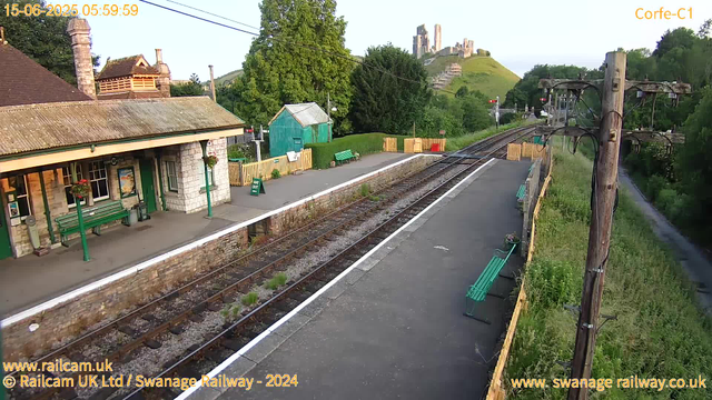 A small railway station platform with a stone and brick building on the left, green benches, a green shed, and flowers hanging in pots. There are several wooden fences painted in light colors along the edge of the platform. Railway tracks run through the center of the image, with a tall wooden utility pole and wires on the right side. In the background, there are lush green trees, a statue or castle ruins on a hill, and a sunny sky with a few clouds. The timestamp shows "15-06-2025 05:59:59."