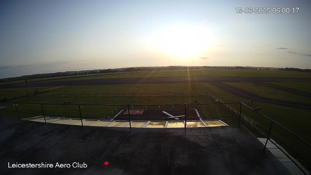 A wide view of an airfield during sunrise, with a grassy area, runways, and airport structures. The sky is mostly clear with the sun low on the horizon, casting a bright glow. The timestamp in the top right corner shows the date as June 15, 2025, and the time as 6:00:17 AM. In the bottom left corner, there is text that reads "Leicestershire Aero Club."
