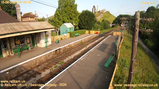 A small train station platform with benches and a building made of stone and brick, featuring hanging flower baskets. There are overhead power lines and a large green tree in the background, along with a grassy hill topped with ruins. The railway tracks run through the station, and there are green benches along the platform, with a small green shed nearby. The scene captures a bright, sunny morning with long shadows.